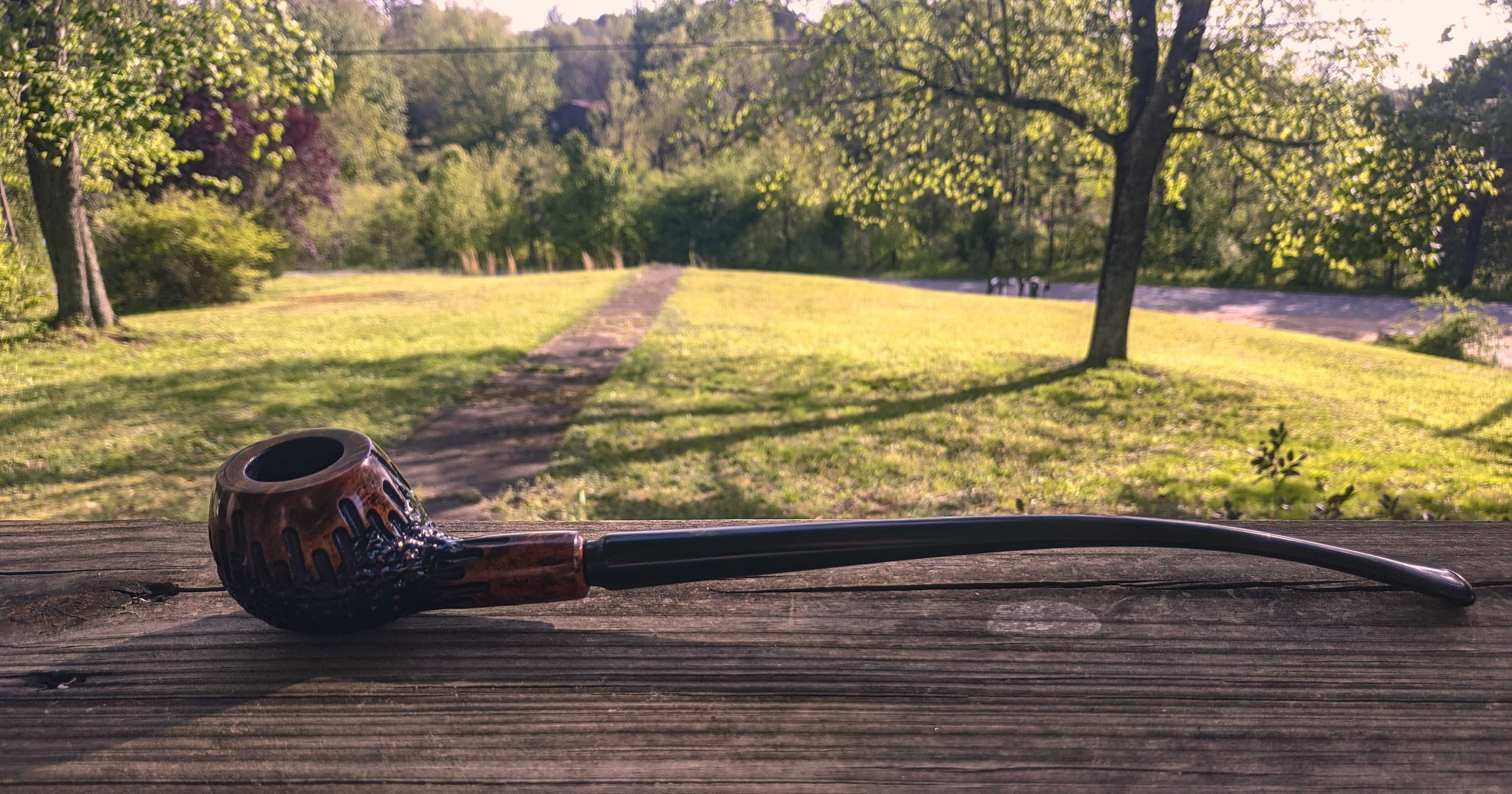 This is also a picture of a pipe on my porch rail. It is significantly
longer at about 10.5 inches or 265 millimetres. The stem has a gentle
curve. The bowl is the same deep brown as the Armellini pipe but the
finish is newer so it shines more. The rustication will be hard to
describe. The rest of the pipe is brown but the rusticated part is
black. The texture is rough but smooth—the edges that might have been
jagged were sanded down. The bottom of the bowl is what’s rusticated;
the top is normal. When held upside down, it almost looks as if the
rusticated part is dripping onto the normal part and the same for the
stem.